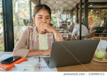 A woman deeply focused on her laptop in a bright cafe, surrounded by documents and a refreshing drink. A woman deeply focused on her laptop in a bright cafe, surrounded by documents and a refreshing drink. 126066313