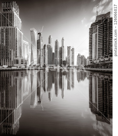 Monochrome city skyline with tall skyscrapers reflecting on calm water under a cloudy sky, Dubai Marina bay UAE 126066817
