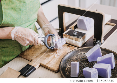 Woman using a vegetable peeler to refine the edges of handmade lavender soap bars on a wooden surface. Sustainable skincare production, home-based business models, self-employment, craft economy Woman using a vegetable peeler to refine the edges of handmade lavender soap bars on a wooden surface. Sustainable skincare production, home-based business models, self-employment, craft economy 126068230