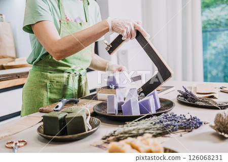 Woman in green apron slicing handmade lavender soap on a worktable with bunches of dried lavender nearby. Clean beauty, natural ingredients, chemical-free soap, small-scale production Woman in green apron slicing handmade lavender soap on a worktable with bunches of dried lavender nearby. Clean beauty, natural ingredients, chemical-free soap, small-scale production 126068231