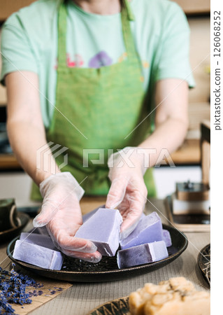 Woman in green apron presenting freshly crafted lavender soap bars on a plate with gloved hands. Handmade wellness, chemical-free skincare, female entrepreneurship, botanical beauty 126068252