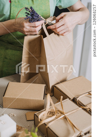 A small business owner ties a Handmade with love tag and lavender sprig to a brown paper bag surrounded by gift boxes. Home-based business, handmade goods 126068257