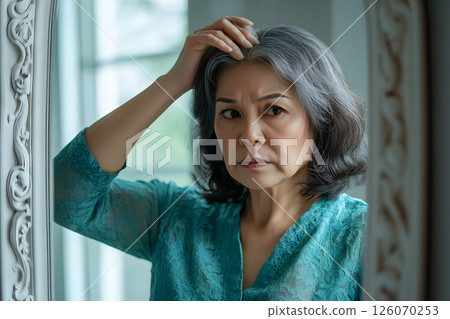 Close-up of realistic facial expression of middle-aged Asian woman worrying about gray hair in front of the mirror 126070253