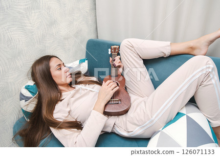 Woman Lounges on a Couch, Wearing Casual Attire, Strumming Ukulele with Focused Expression. Woman Lounges on a Couch, Wearing Casual Attire, Strumming Ukulele with Focused Expression. 126071133