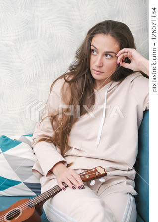 Woman with Long Hair Sits Thoughtfully on Couch, Holding Ukulele. She Gazes into the Distance, Creating Relaxed Atmosphere. 126071134