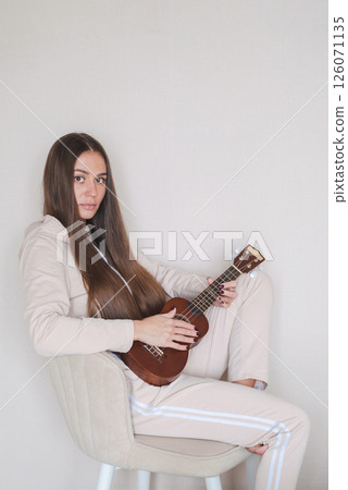 Woman with Long Hair Sits in Comfortable Chair While Skillfully Playing the Ukulele. 126071135