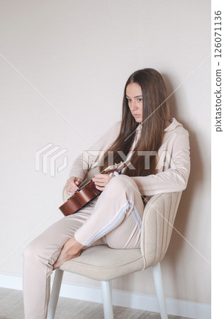 Woman Sits Comfortably on a Chair, Playing Ukulele Quietly. She Wears Casual Clothes and Seems Lost in Her Music. 126071136