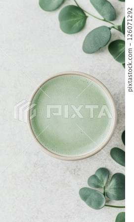A cup of frothy green matcha latte sits on a light grey surface, adorned with fresh eucalyptus leaves A cup of frothy green matcha latte sits on a light grey surface, adorned with fresh eucalyptus leaves 126071292