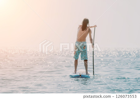 Man Paddleboarding Ocean Sunset - Man paddling on a surfboard on a calm ocean at sunset. Man Paddleboarding Ocean Sunset - Man paddling on a surfboard on a calm ocean at sunset. 126073553