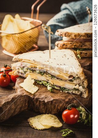 Delicious sandwich stacked with layers of fresh vegetables, meats, and cheese, served on a rustic wooden board with crispy chips and cherry tomatoes, showcasing appetizing food presentation 126073626