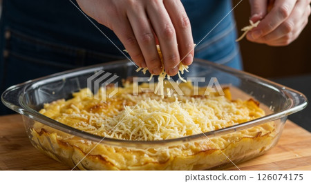 Woman sprinkling grated cheese on potato gratin in glass dish on kitchen counter before baking 126074175