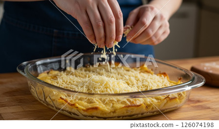Woman sprinkling grated cheese on potato gratin in glass dish on kitchen counter before baking Woman sprinkling grated cheese on potato gratin in glass dish on kitchen counter before baking 126074198