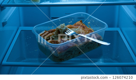 Portion of leftover Greek pastitsio in plastic container with fork inside lit blue refrigerator Portion of leftover Greek pastitsio in plastic container with fork inside lit blue refrigerator 126074210