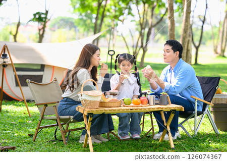 Cheerful family camping outdoors, sharing snacks and spending quality time near tent in the park. Cheerful family camping outdoors, sharing snacks and spending quality time near tent in the park. 126074367
