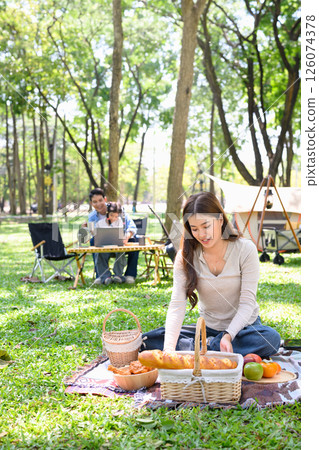 Woman arranging food for a picnic while her family enjoys time at a forest campsite Woman arranging food for a picnic while her family enjoys time at a forest campsite 126074378