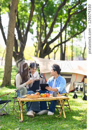 Happy family enjoying a camping trip sitting by the tent with food and browsing on a tablet Happy family enjoying a camping trip sitting by the tent with food and browsing on a tablet 126074380