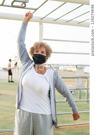 Elderly woman in protective mask doing fitness on gym equipment on beach Elderly woman in protective mask doing fitness on gym equipment on beach 126074696