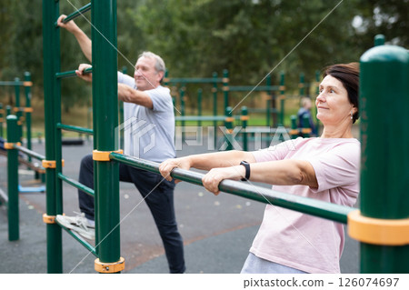 Elderly man and woman doing exercises on horizontal bars at outdoor sports ground Elderly man and woman doing exercises on horizontal bars at outdoor sports ground 126074697