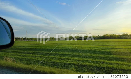 Stunning view of the green meadow from the side window of a moving car. car mirror in the frame, and car is moving at speed along a picturesque rural area in the evening. Stunning view of the green meadow from the side window of a moving car. car mirror in the frame, and car is moving at speed along a picturesque rural area in the evening. 126075350