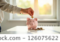 hand placing coin in piggy bank by a sunny window, depicting savings growth, ideal for financial planning, personal savings, and investment strategies 126075622
