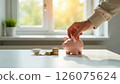 hand placing a coin in a piggy bank by a sunny window, depicting savings growth, ideal for financial planning, personal savings, and investment strategies 126075624