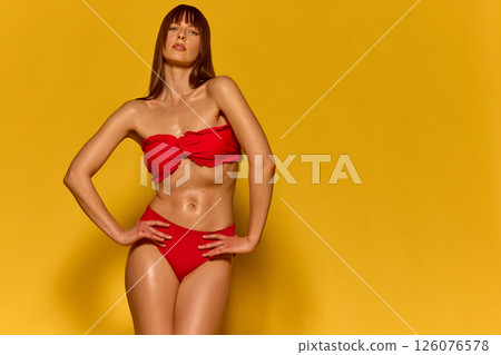 Elegant female model with long hair and fringe posing in red bikini top with expressive arm posture against yellow studio background. 126076578