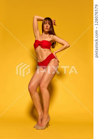 Slim woman wearing red retro bikini with ruffles and nude heels posing elegantly with hand on head against yellow background. 126076579