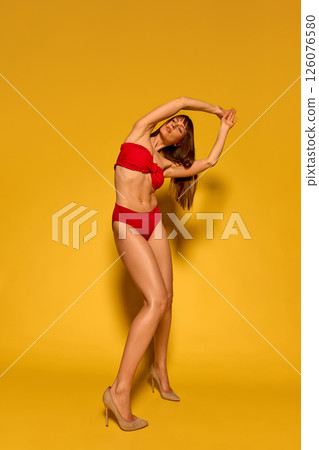 Toned female body in red ruffled bikini captured in twisted motion with graceful curved arms against yellow studio background. 126076580