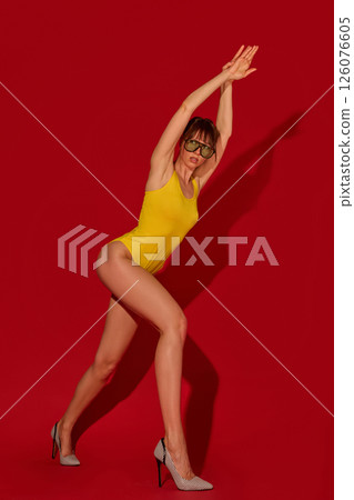 Fit woman with long legs in yellow swimsuit extends arms above head in motion wearing green sunglasses and heels against red studio background 126076605