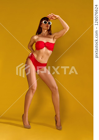 Woman in red bikini wearing white sunglasses posing creatively against yellow background embracing retro holiday style and vibrant summer expression. 126076624