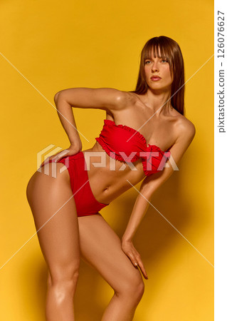 Close up side view of woman in red bikini on yellow background showing summer elegance retro charm and feminine posture in beach fashion. 126076627