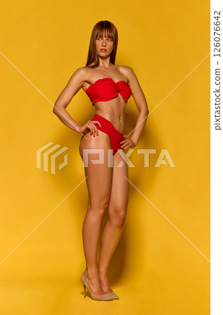 Full body portrait of fit woman standing confidently in red retro bikini and beige heels showing toned muscles against yellow background. 126076642
