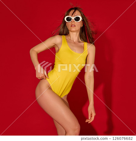 Woman in yellow bodysuit places hand on waist looking up with sunglasses showing sleek lines and empowered pose against red background 126076682