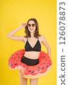 Stylish young woman in black bikini holding pink donut pool float and wearing sunglasses on yellow background, smiling and posing playfully for summer travel, swimwear, or vacation-themed concepts 126078873