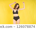 Energetic young woman dancing joyfully in black bikini against vibrant yellow studio background, expressing fun, freedom, and confidence for fashion, lifestyle, or health themes 126078874