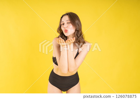 Confident young brunette woman in black bikini blowing a kiss while smiling in studio with yellow background, expressing fun playful energy for beauty, fashion, or lifestyle concepts 126078875