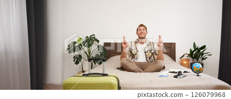 Portrait of young man meditating on a bed after packing suitcase for summer holiday, sitting with crossed legs in zen, nirvana pose, relaxing, hoping for something Portrait of young man meditating on a bed after packing suitcase for summer holiday, sitting with crossed legs in zen, nirvana pose, relaxing, hoping for something 126079968