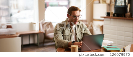 Portrait of man with serious face, sitting in cafe, wearing glasses, working on laptop, looking concentrated, doing freelance project Portrait of man with serious face, sitting in cafe, wearing glasses, working on laptop, looking concentrated, doing freelance project 126079970