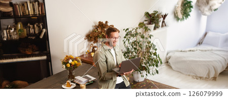 Vertical portrait of handsome happy man, manager or CEO of company, sitting on table and holding laptop, laughing, video chatting, has an online meeting with a team, standing in an office 126079999