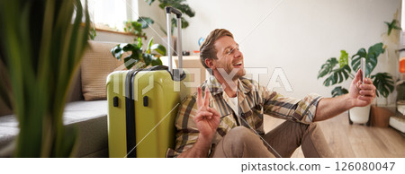 Portrait of smiling handsome man, shows peace sign, takes selfies on smartphone, sits with suitcase, goes on holiday Portrait of smiling handsome man, shows peace sign, takes selfies on smartphone, sits with suitcase, goes on holiday 126080047