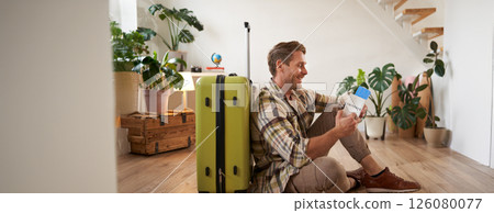 Portrait of handsome young man, tourist sitting with his suitcase, holding passport and flight tickets, going on holiday Portrait of handsome young man, tourist sitting with his suitcase, holding passport and flight tickets, going on holiday 126080077