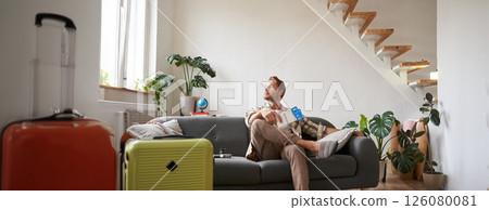 Portrait of handsome young man, sits with suitcase in living room, holds two plane tickets, going on vacation, waiting for taxi, looking outside window 126080081