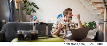 Upbeat man, tourist with two tickets, makes a fist pump, has laptop opened, goes on vacation, excited about holiday Upbeat man, tourist with two tickets, makes a fist pump, has laptop opened, goes on vacation, excited about holiday 126080083