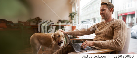 Handsome young man working in cafe with a dog, sitting on chair and using laptop, petting his golden retriever in animal-friendly co-working space Handsome young man working in cafe with a dog, sitting on chair and using laptop, petting his golden retriever in animal-friendly co-working space 126080099
