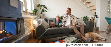 Portrait of young man sitting on sofa with notebook, has dog lying next to him, packing suitcase for summer vacation 126080100