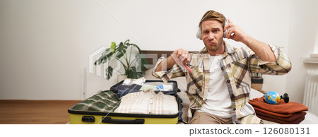 Portrait of disappointed man in headphones, listen to music, shows thumbs down, dislike hand gesture, sits in a hotel with unpacked suitcase Portrait of disappointed man in headphones, listen to music, shows thumbs down, dislike hand gesture, sits in a hotel with unpacked suitcase 126080131