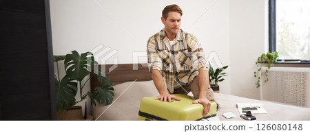 Portrait of young man, tourist packs his bag, closing full suitcase, trying to push the luggage inside to zip the bag, sitting on bed in bedroom 126080148