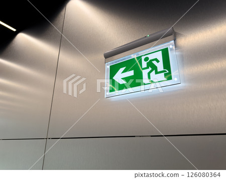 Glowing green emergency exit sign with arrow and running person symbol on brushed metal wall. Evacuation indicator in modern public building. Concept of alert, escape route and safety compliance Glowing green emergency exit sign with arrow and running person symbol on brushed metal wall. Evacuation indicator in modern public building. Concept of alert, escape route and safety compliance 126080364