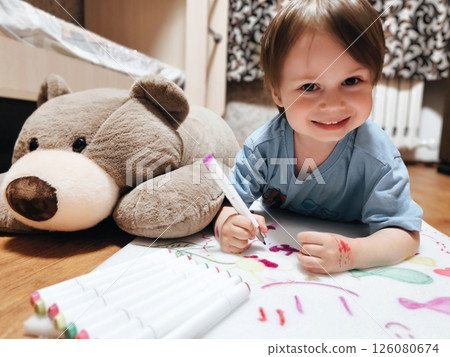 Little boy draws with colored markers. child draws in his room lying on the floor. A teddy bear next to the baby. 126080674
