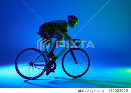 Side view image of male cyclist in full gear, riding bicycle, training against blue background with green neon light. 126083056